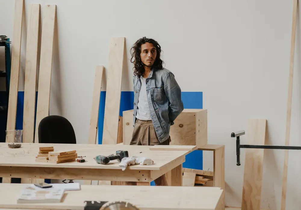 Un homme debout dans un atelier de menuiserie, entre des panneaux de bois appuyés au mur et un établi où sont posés une perceuse et des chutes