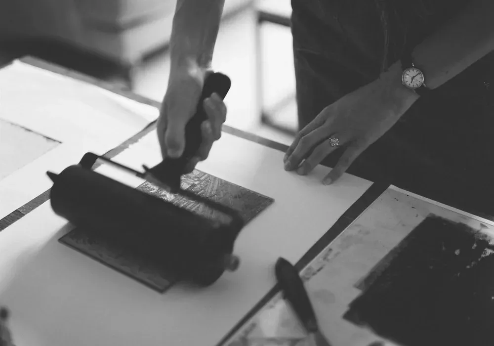 Une personne encre une plaque de gravure au rouleau sur une table d'atelier, une montre au poignet