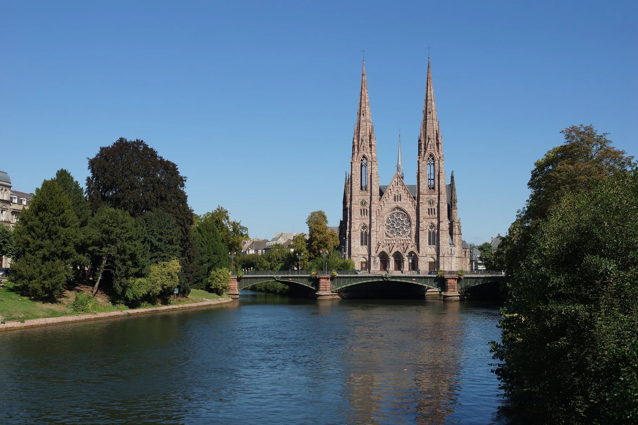 L’église Saint-Paul de Strasbourg au bord de l’Ill, sous un ciel bleu, avec un pont et les arbres des quais